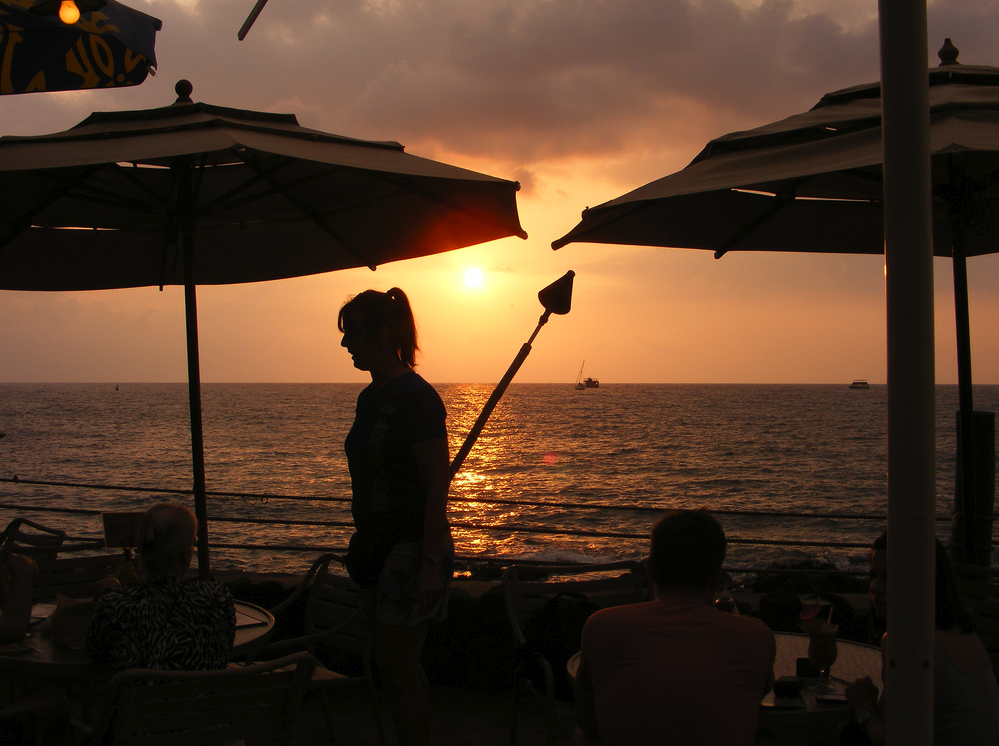 People dining at an outdoor restaurant by the beach at sunset with umbrellas shading the tables